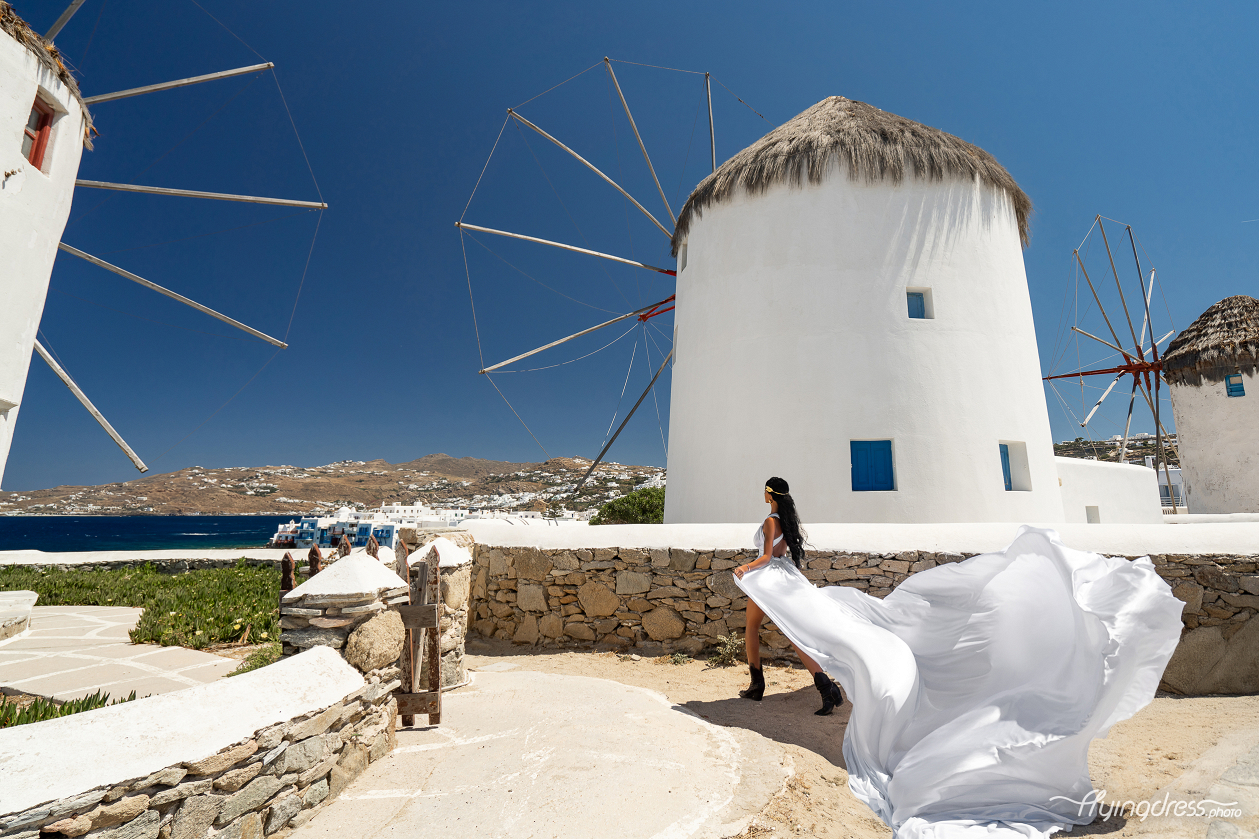 A woman in a flowing white dress poses next to a traditional Mykonos windmill, with the deep blue sky and sea providing a stunning backdrop.